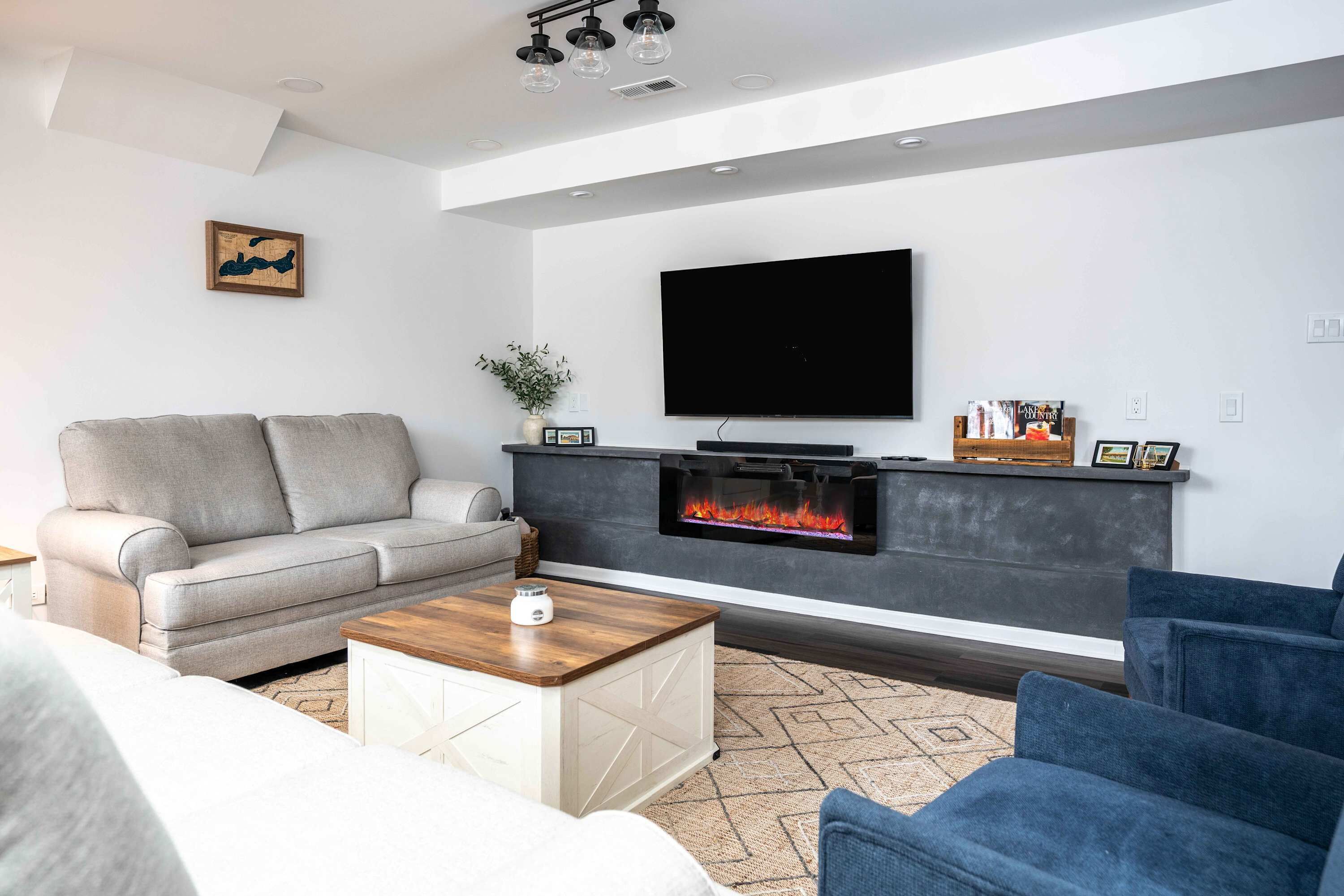 Una sala de estar moderna con un sofá gris, un sillón azul, una mesa de centro, una chimenea y un televisor de pantalla plana montado en la pared.