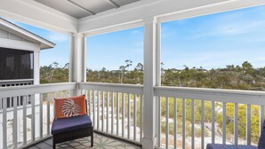 A covered outdoor area with a white railing, a bench with a cushion, and a view of the beach and trees.