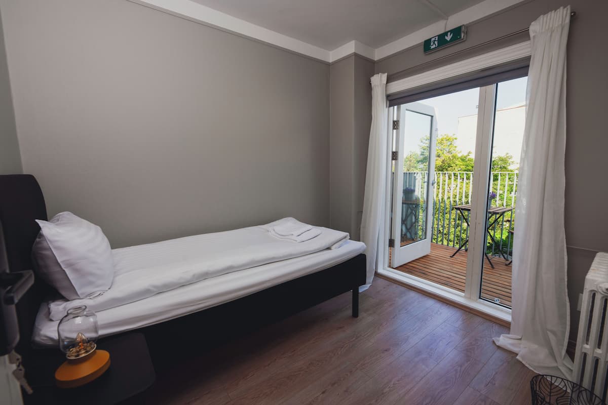 A single bed with a white mattress and pillow, a small bedside table with a glass container, and a view of a wooden deck through a sliding glass door.