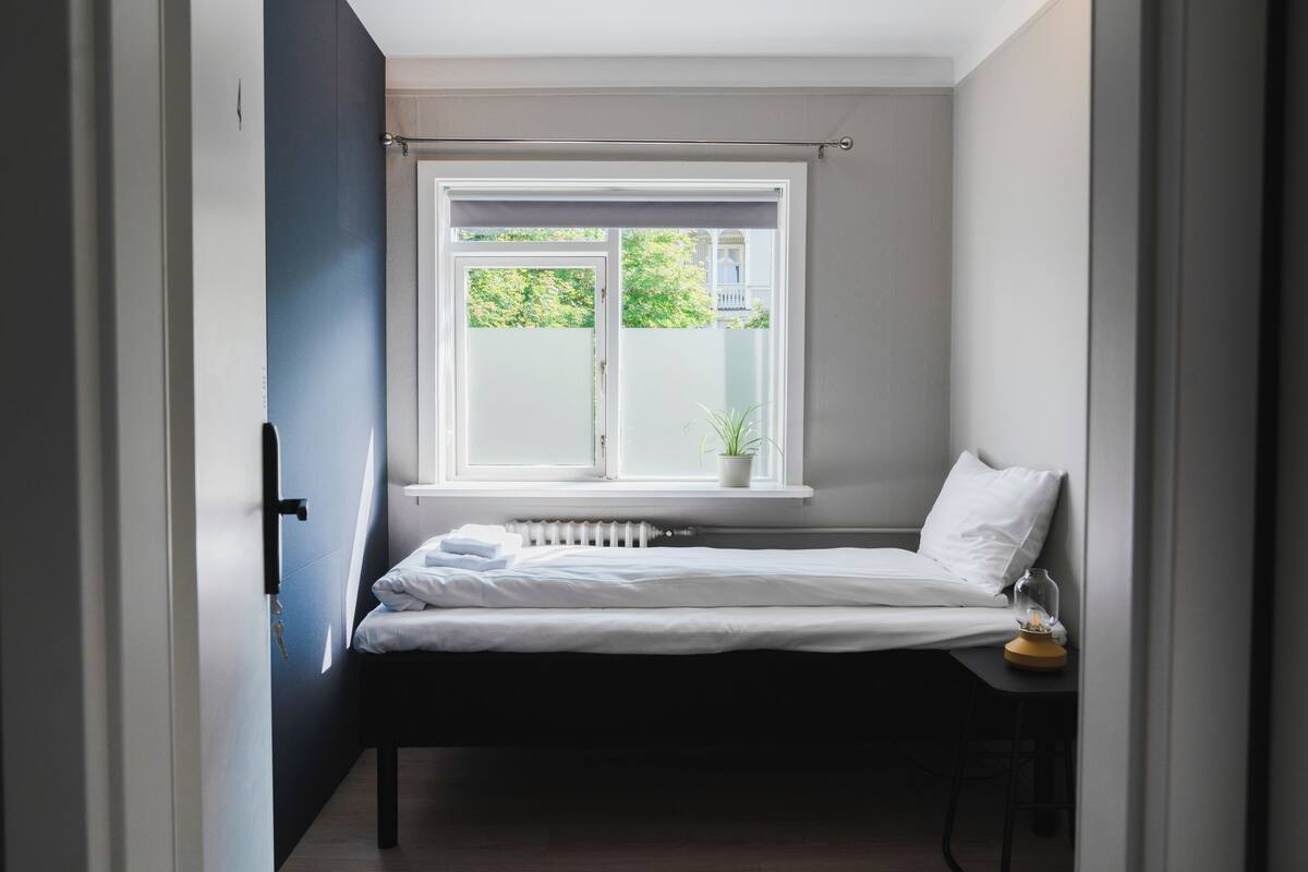 A bedroom with a bed, a window with a view of greenery, a radiator, and a small table with a lamp.
