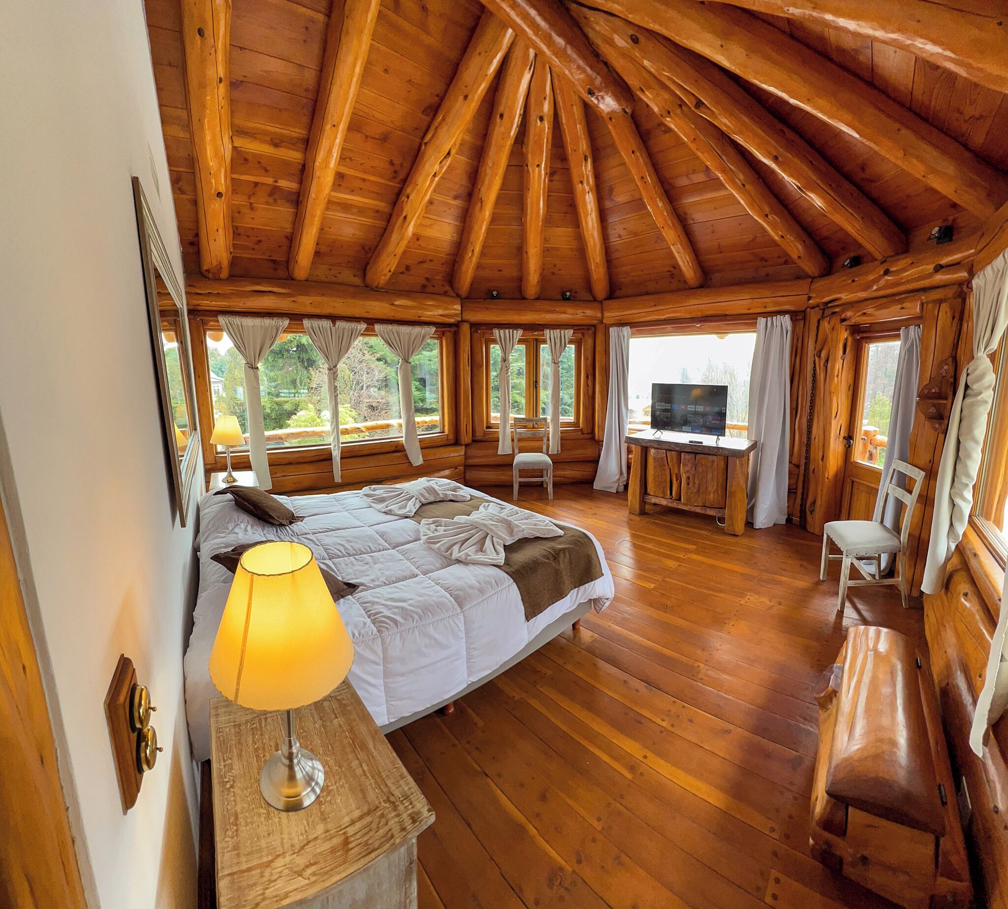 A wooden cabin bedroom with a bed, bedside lamp, and a desk with a TV.