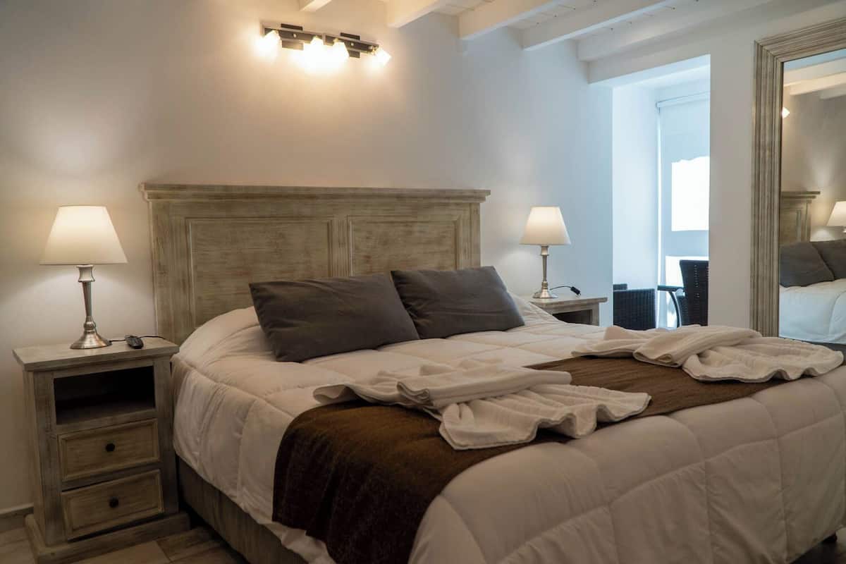 Una cama bien hecha con cabecera de madera, dos mesitas de noche con lámparas y un espejo grande en la pared.
