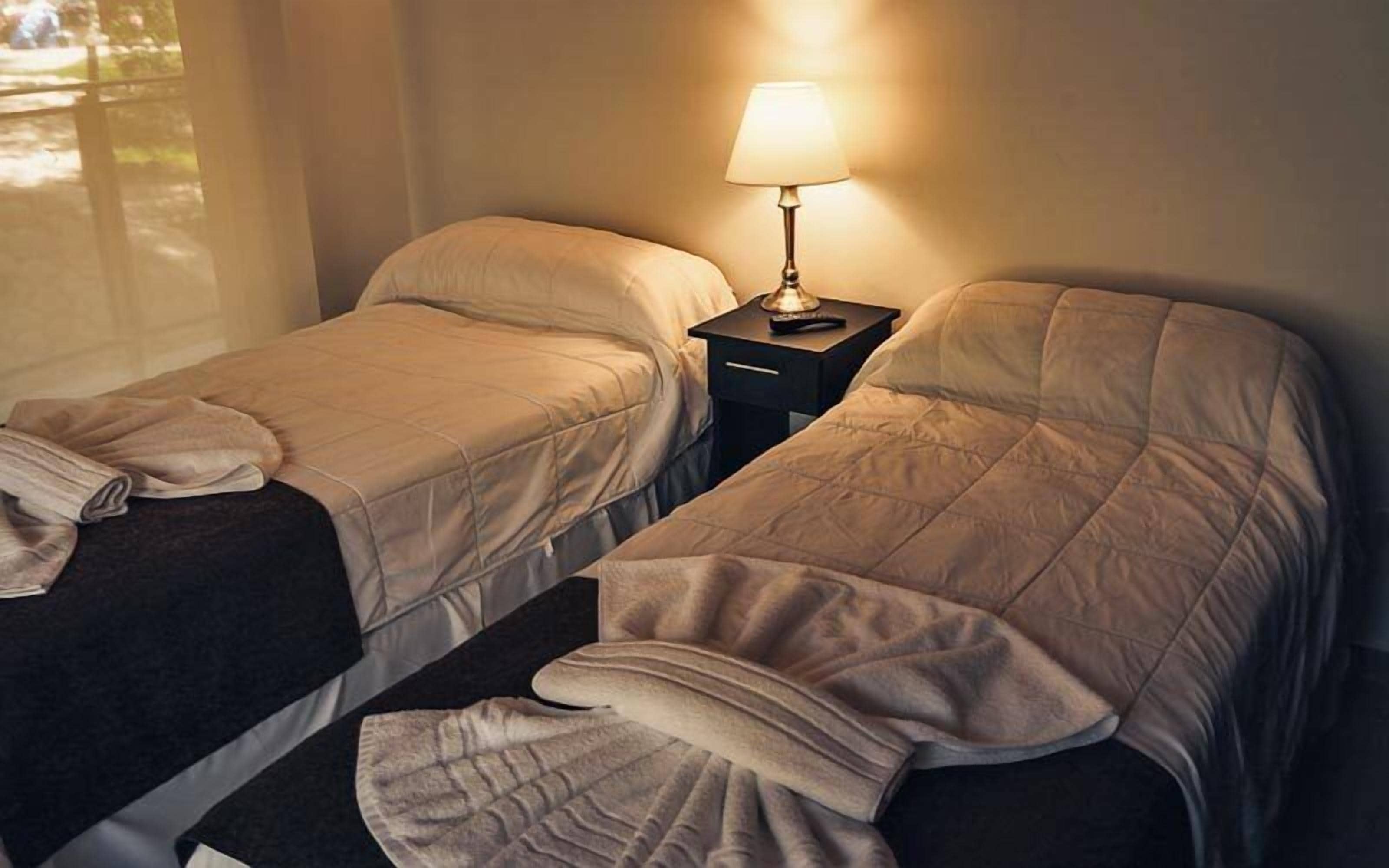 Two single beds with beige bedding, a nightstand with a lamp, and a window with natural light.