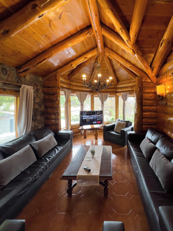 A log cabin interior with a stone wall, wooden ceiling, and a large TV screen. - Rotui Luxury Village (San Martín de los Andes)