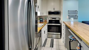 Moderni keittiö, jossa on ruostumattomasta teräksestä valmistetut kodinkoneet, valkoiset kaapit, mikroaaltouuni ja musta uuni. Keittiössä on puinen työtaso ja värikäs istuinosaa sisältävä jakkara.