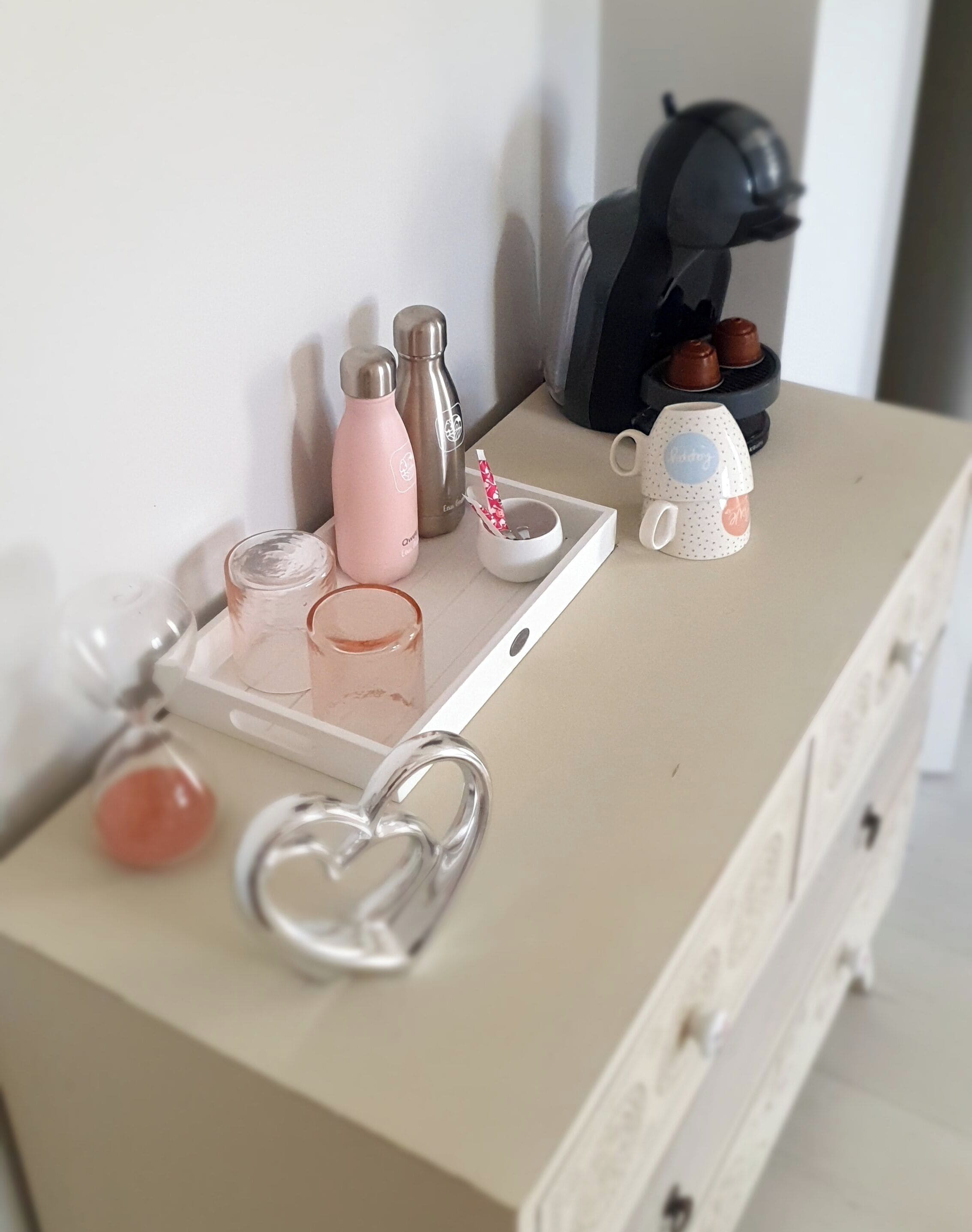 Et lille, pænt arrangeret sminkebord med en hvid skuffe, en bakke med forskellige genstande og en dekorativ figur.