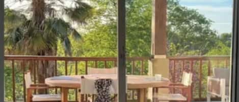 A balcony with a wooden table and chairs, a patterned rug, and a view of palm trees and greenery.