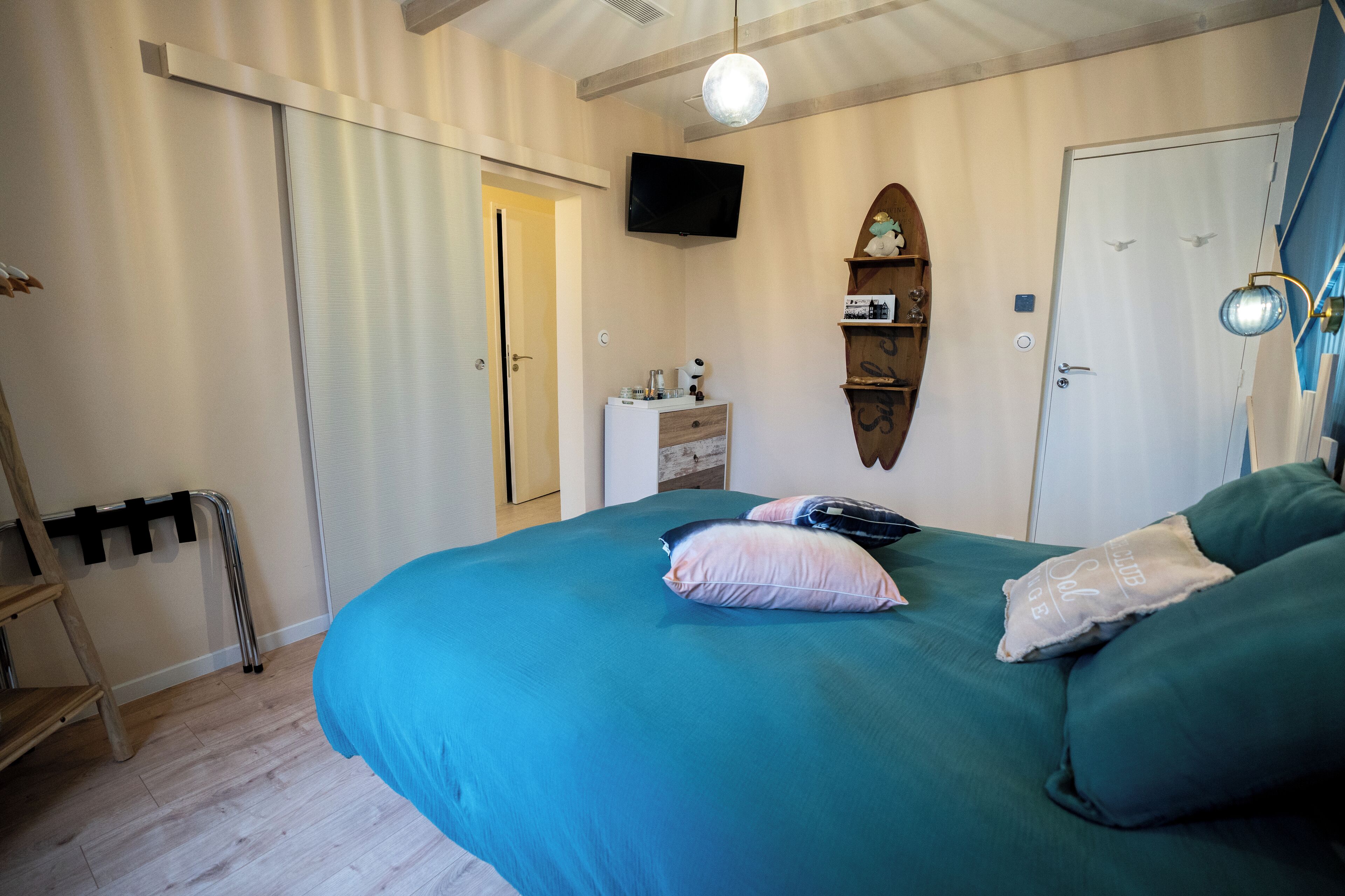 A bedroom with a teal bedspread, a wooden ladder, a white dresser, a television mounted on the wall, and a door with a bird decal.