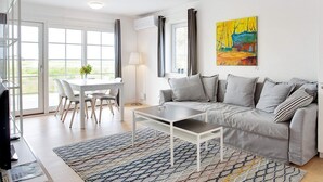 A modern living room with a grey sofa, a coffee table, a dining table with chairs, and a colorful painting on the wall. - Nordic Pearl: Premium Holiday Home in Laeso (Læsø)