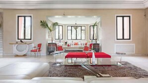 Un salon moderne avec un ensemble de canapés, une table basse et un pouf rouge. On y trouve également deux fauteuils, une table d’appoint et une plante.