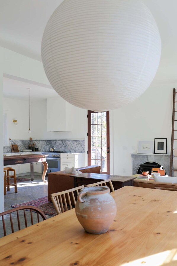 Une cuisine moderne avec un plan de travail en marbre, un grand luminaire suspendu blanc et une table à manger en bois rustique agrémentée d’un centre de table en poterie.