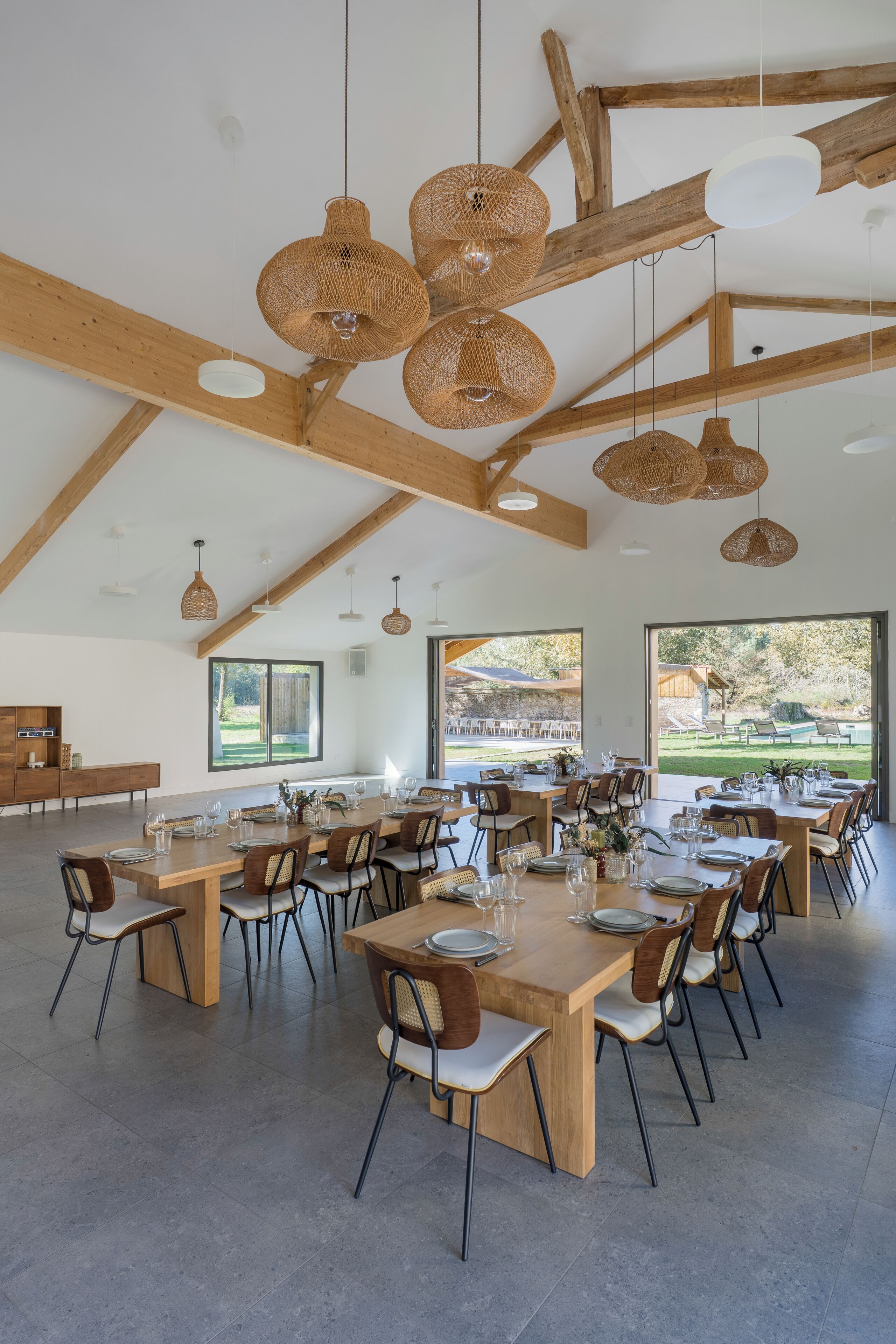 Un comedor amplio con varias mesas y sillas de madera, ventanas grandes que ofrecen vistas al exterior y lámparas colgantes tejidas.