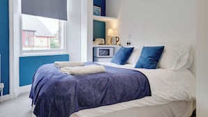 A bedroom with a large bed, a bedside table with a lamp, and a view of a red building outside. - Homesly Guest Rooms - 1 Bed Room Apt. (Berwick-upon-Tweed)