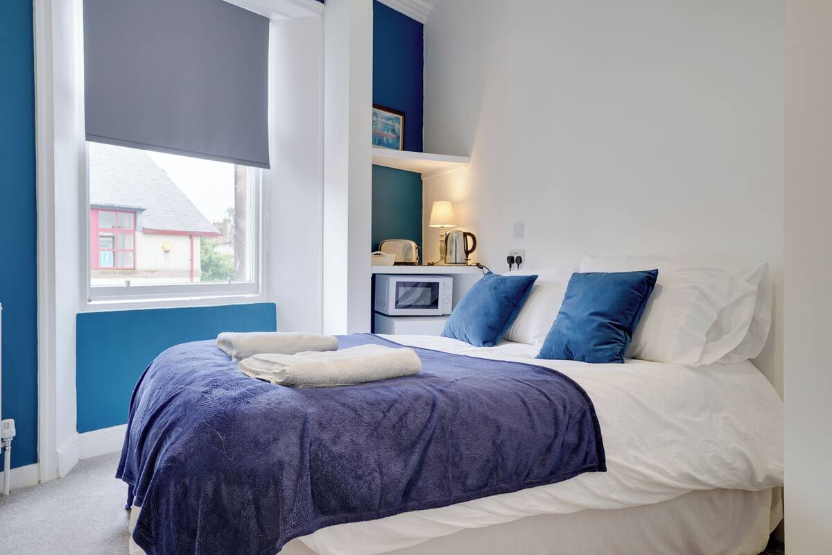 A bedroom with a large bed, a bedside table with a lamp, and a view of a red building outside.