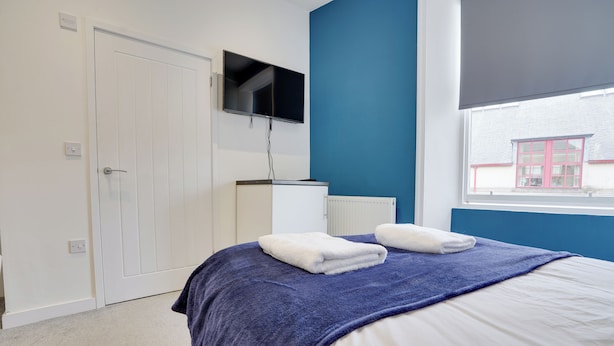A bedroom with a bed, a TV mounted on the wall, a window, and a radiator. - Homesly Guest Rooms - 1 Bed Room Apt. (Berwick-upon-Tweed)