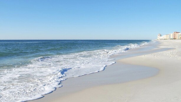 A beach with a clear sky, gentle waves, and a row of buildings in the background. - Bay Breeze Retreat- So Close To All The Fun! Game Room! XL Home! (Orange Beach)