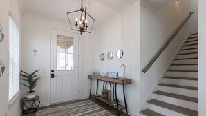 A hallway with a wooden floor, a striped rug, a console table with decorative items, a chandelier, a staircase, and a potted plant. - Bay Breeze Retreat- So Close To All The Fun! Game Room! XL Home! (Orange Beach)