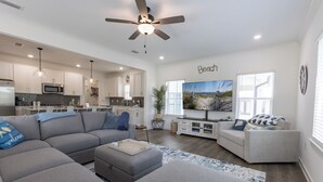 A modern living room with a grey sectional sofa, a grey armchair, and a coffee table. There is a flat-screen TV mounted on the wall, a ceiling fan, and a kitchen area with white cabinets and a microwave. - Bay Breeze Retreat- So Close To All The Fun! Game Room! XL Home! (Orange Beach)