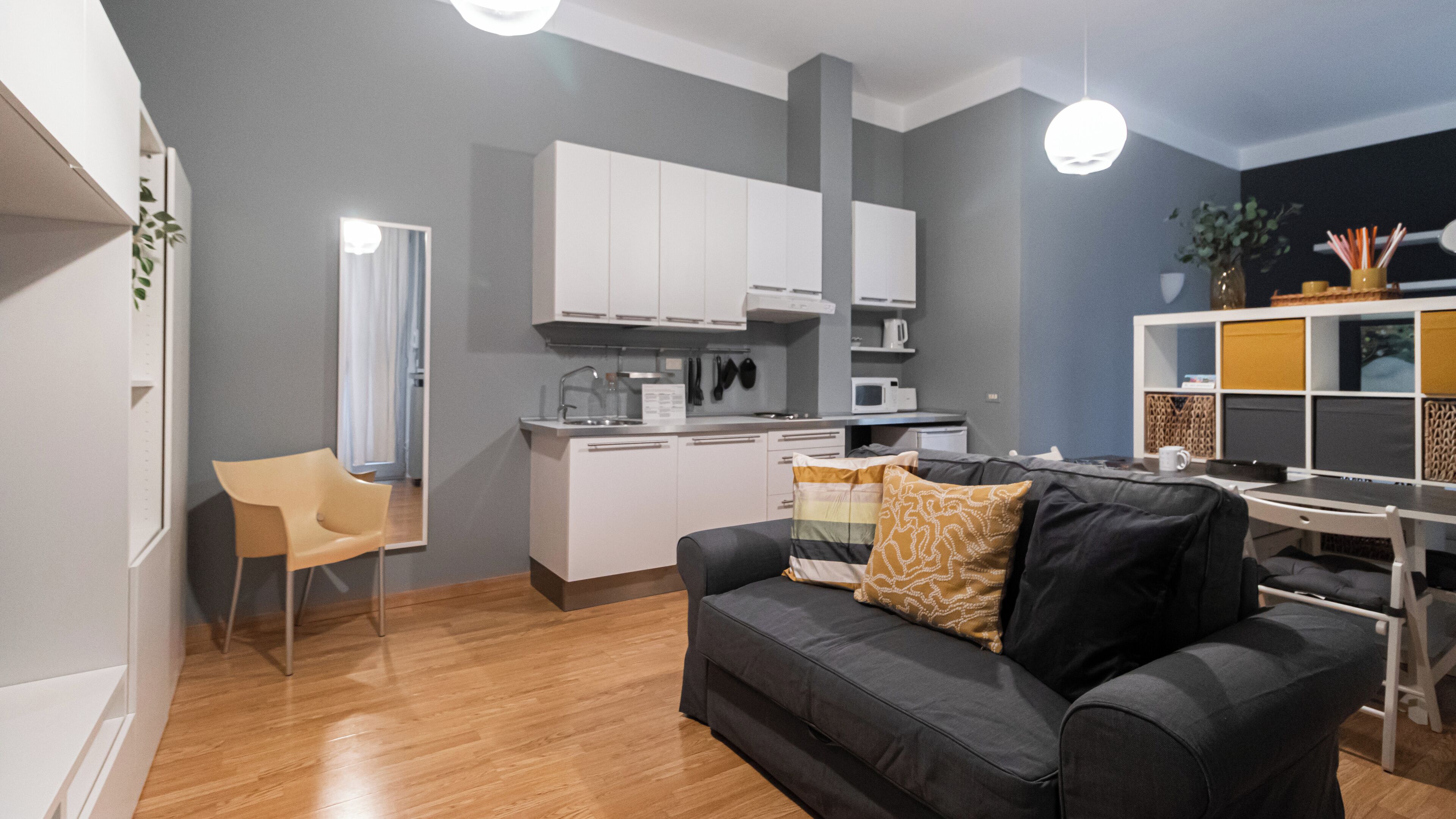 A modern living room with a black sofa, a wooden floor, and a kitchen in the background.