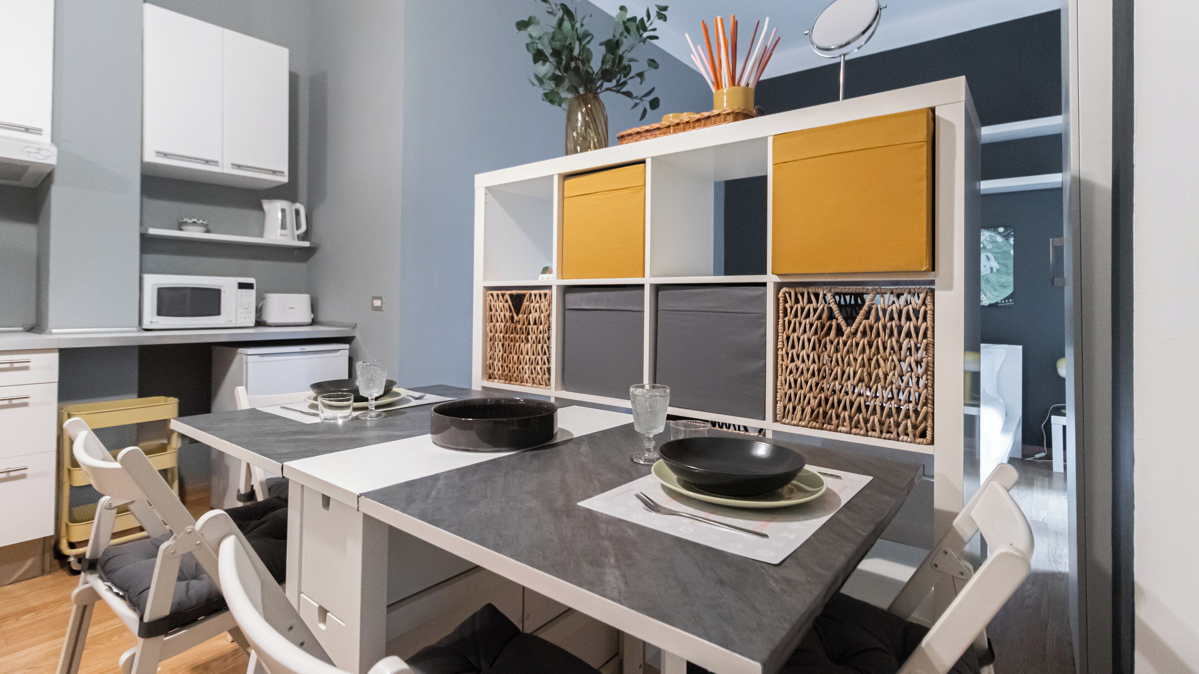A modern kitchen with white cabinets, a gray countertop, and a yellow accent wall. There is a microwave, a coffee maker, and a vase with a plant on the counter.