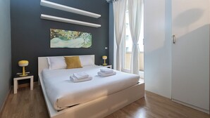 Egy modern szállodai szoba, amelyben egy nagy ágy, fehér ágynemű, két éjjeliszemélyke lámpákkal, egy kép a falon és egy ablakból nyíló kilátás található.