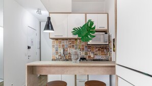 Une cuisine avec un plan de travail blanc, un four à micro-ondes, une grande plante verte et deux tabourets en bois.