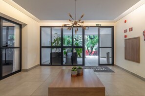 A modern lobby with a wooden reception desk, a unique chandelier, glass doors leading to an outdoor area with plants, and a fire safety sign on the wall.
