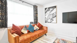 A living room with a brown sofa, patterned curtains, a black abstract painting, and a television. - Host & Stay | Hazelnut House (Launceston)