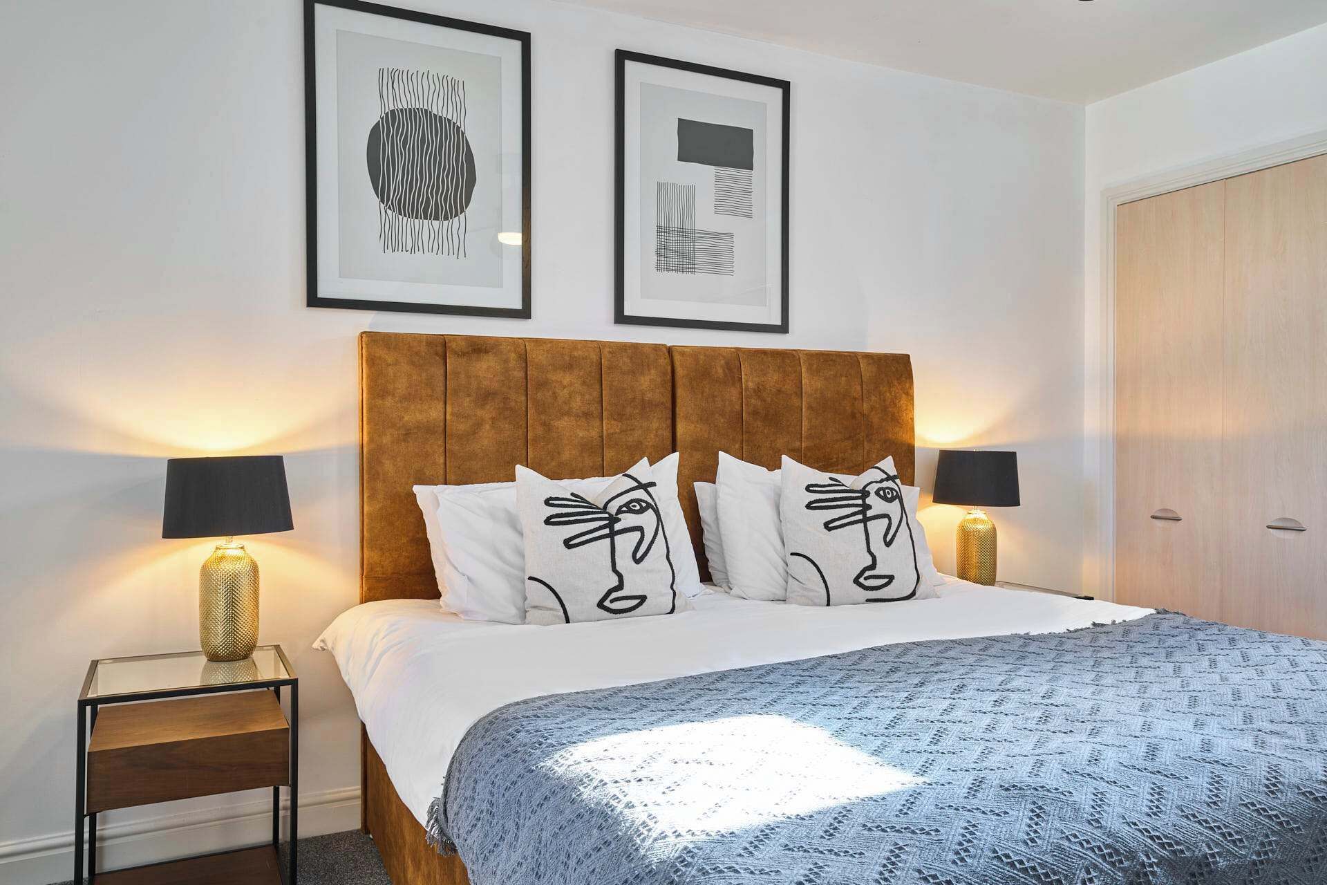 A bedroom with a wooden headboard, white bedding, and patterned pillows. There are two framed artworks on the wall and a bedside table with lamps.