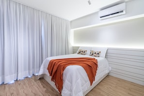 A bedroom with a bed, white curtains, an air conditioning unit, and a bedside table. - Complete Studio in Perdizes: PUC one step away, with Swimming Pool and Fitness (São Paulo)