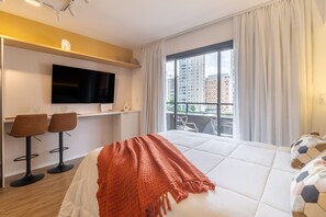 Habitación de hotel con una cama grande, un televisor montado en la pared, dos taburetes de bar, un balcón con vistas a edificios y una ventana con cortinas.