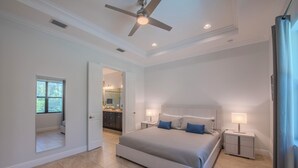 Une chambre spacieuse avec un grand lit, deux tables de chevet avec des lampes, un ventilateur de plafond et une vue sur la salle de bain par une porte ouverte.