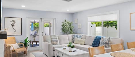 A modern living room with a sofa, coffee table, and chairs. There is a door leading to an outdoor area with plants.