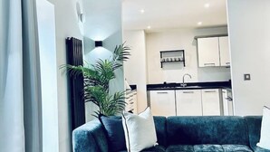 A modern living room with a dark green sofa, a white cushion, and a large plant. A kitchen with white cabinets and a black countertop is visible in the background.