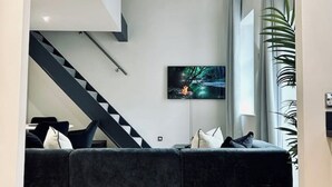 A modern living room with a dark grey sofa, a staircase with black and white steps, and a large window with a plant on the right.