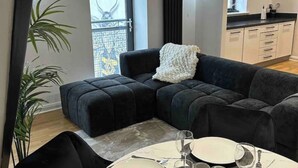 A modern living room with a dark-colored sectional sofa, a white throw blanket, and a dining table set for two with plates, glasses, and cutlery.