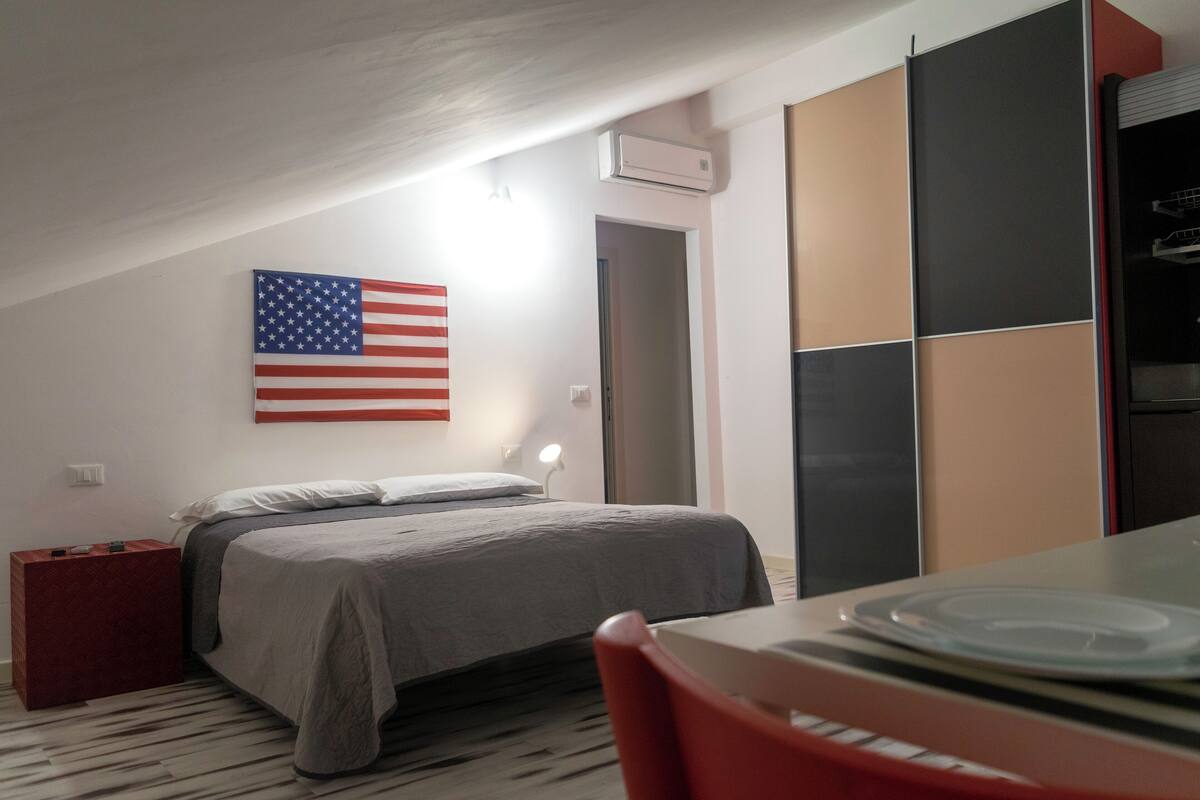Une chambre à coucher avec un lit, une table sur laquelle se trouve une assiette, une chaise rouge, un drapeau américain et une porte.