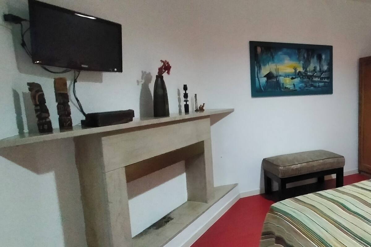 Une chambre à coucher avec un lit, un téléviseur fixé au mur, un tableau, une armoire en bois et un petit tabouret.