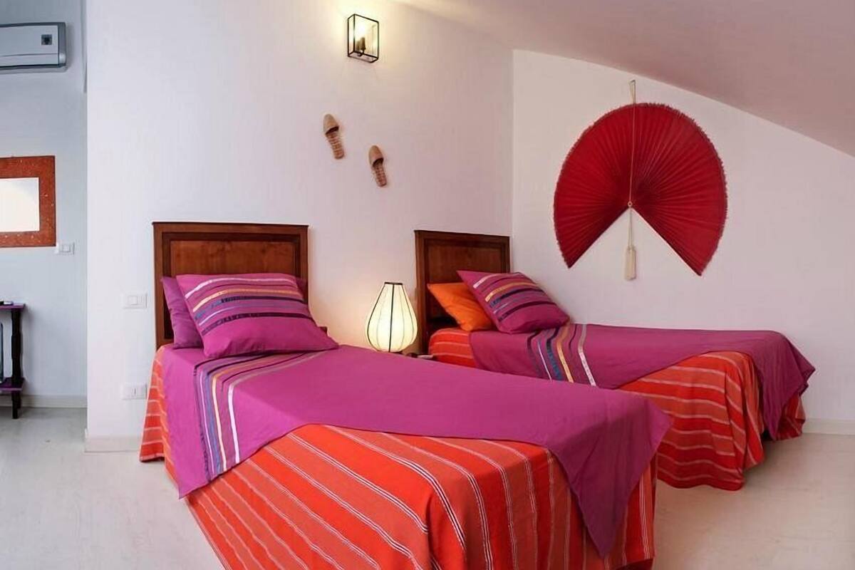 Une chambre d’hôtel avec deux lits, chacun doté de draps et d’oreillers à rayures. On y trouve un ventilateur fixé au mur et une petite table avec une lampe.