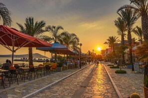 A paved walkway lined with outdoor seating and palm trees, leading to a sunset view. - Marina's Cozy Escape-Sea & Sun Getaway (Romanos)