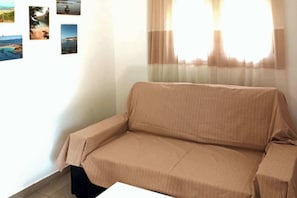 A beige sofa in a room with a window covered by sheer curtains, a tiled floor, and a wall with several framed pictures. - Marina's Cozy Escape-Sea & Sun Getaway (Romanos)