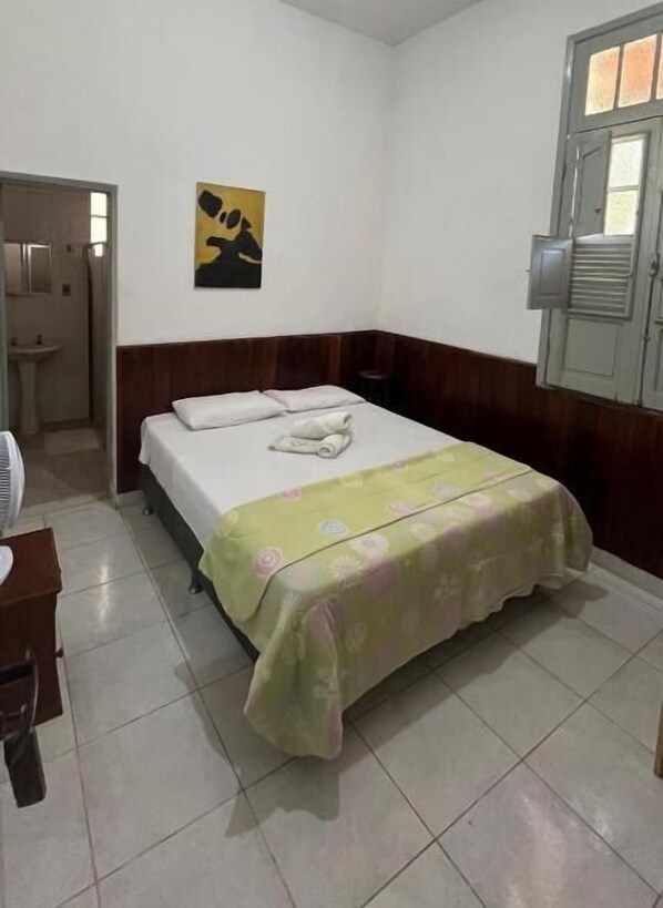 A hotel room with a bed, a bedside table, a chair, a window, and a bathroom visible through an open door. - Hotel Sacra Família  (Engenheiro Paulo de Frontin)