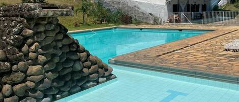 Una piscina con un muro de piedra, un edificio blanco con techo rojo y exuberante vegetación al fondo.