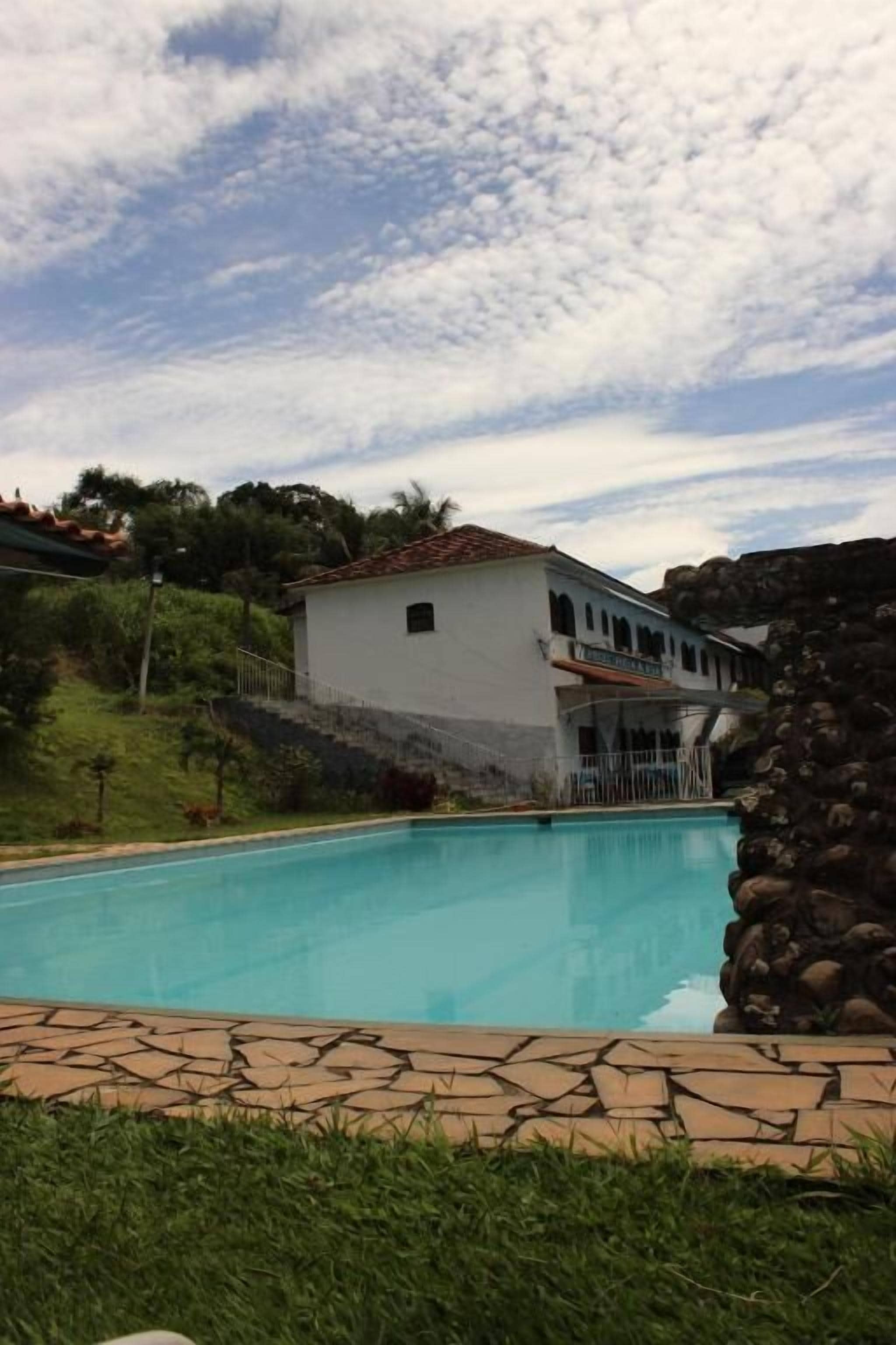 A swimming pool with a stone border, a white building with a red roof in the background, and a grassy area.