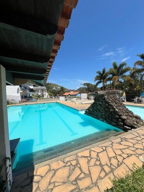 A swimming pool with a stone wall and a view of houses and palm trees in the background. - Hotel Sacra Família  (Engenheiro Paulo de Frontin)