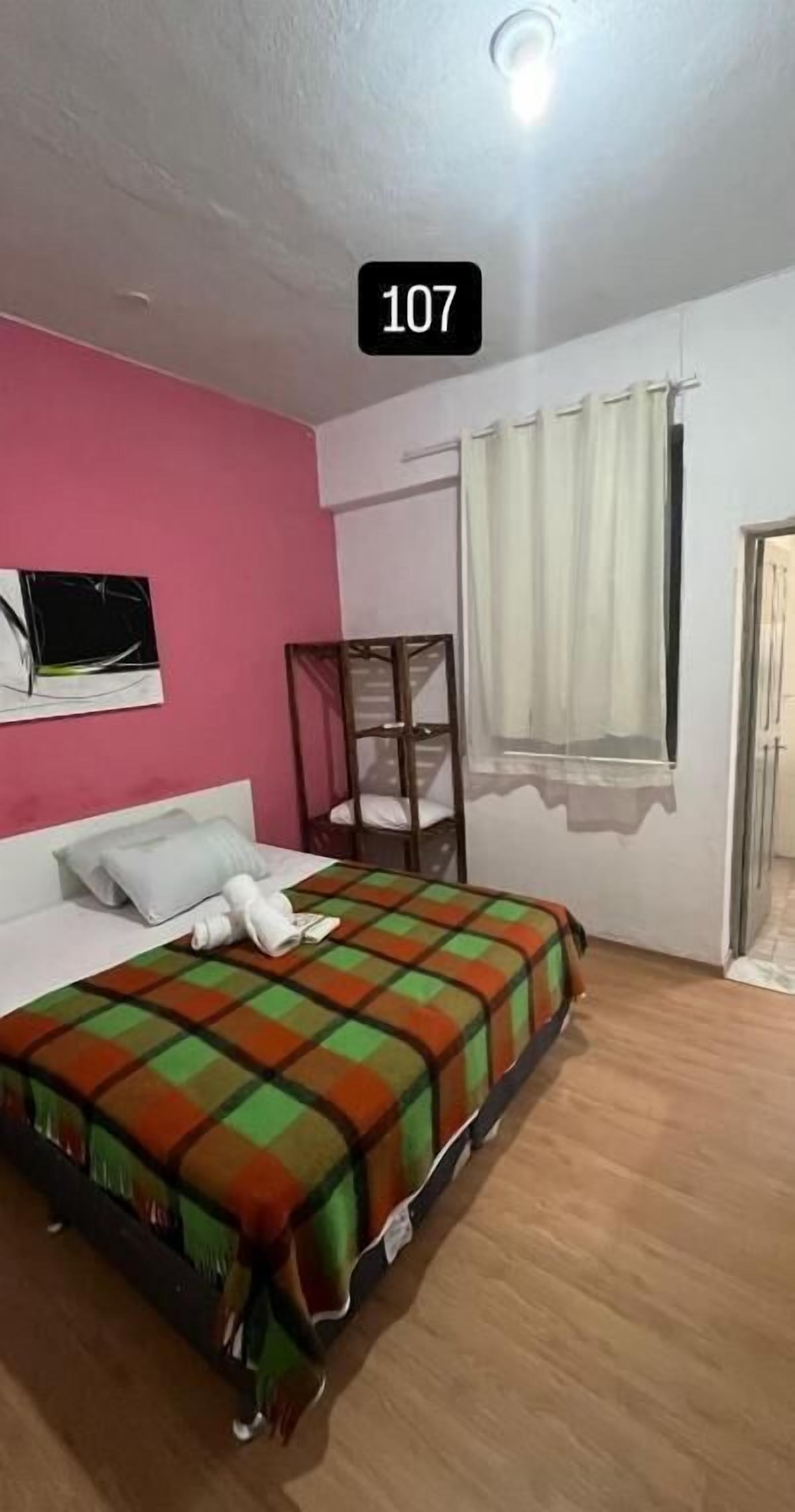Quarto de hotel com cama, janela com cortinas, mesa de cabeceira e televisão fixada na parede.
