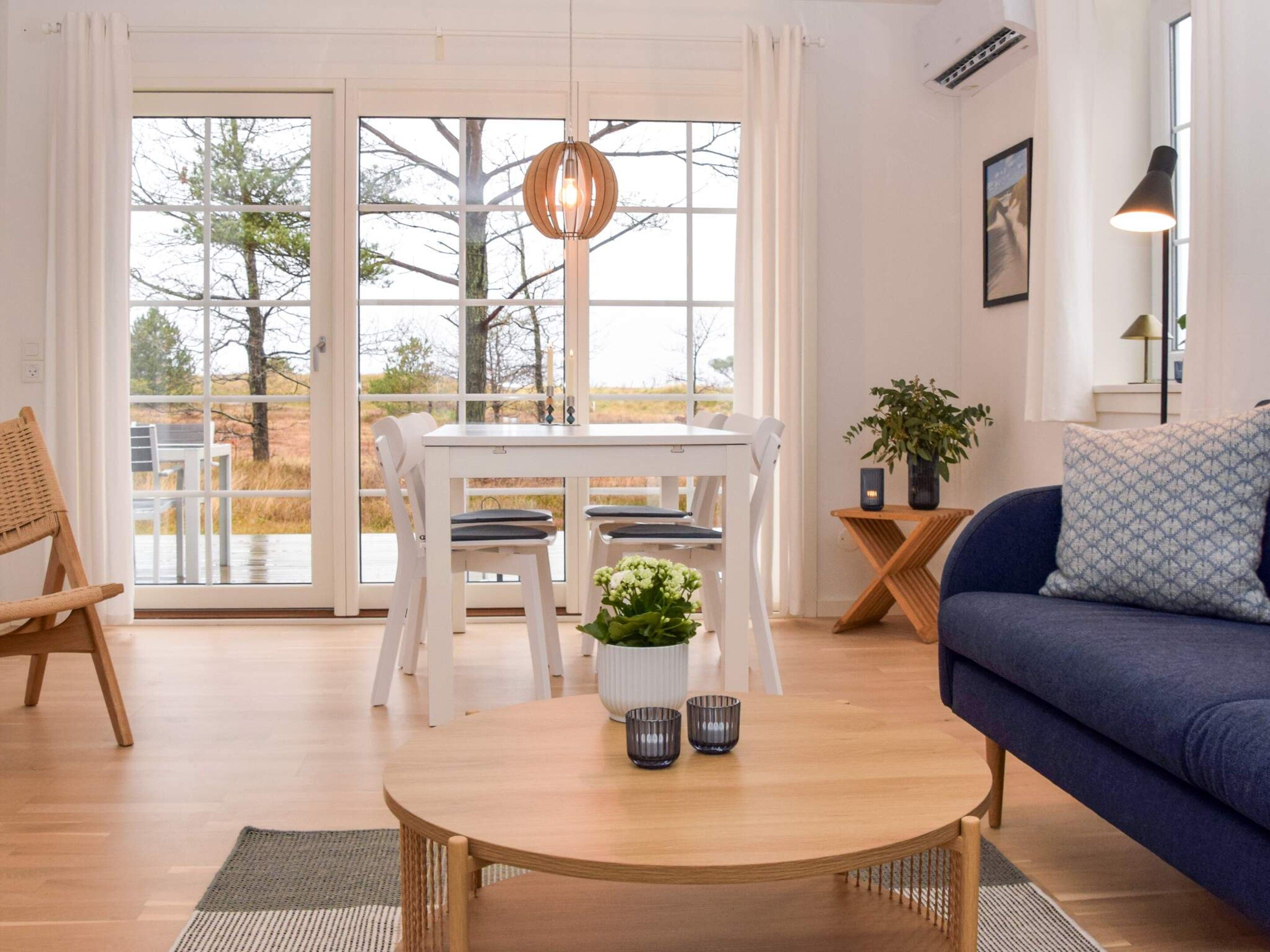 Uma sala de estar moderna com área de jantar, sofá, mesa de centro e vista para o exterior através de grandes janelas.