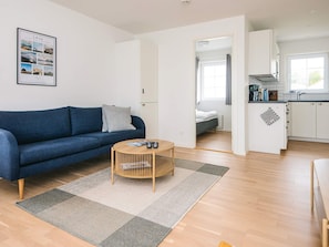 A modern living room with a blue sofa, a wooden coffee table, and a kitchen area with white cabinets and a window. - 4 Star Holiday Home in Laeso (Læsø)