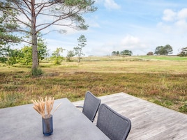 Un espacio exterior para comer con mesa y sillas, un florero con plantas secas, y vistas a un campo cubierto de césped y árboles.