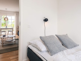 Quarto com cama, almofadas, mesa de cabeceira, abajur e vista para uma área de refeições com mesa e cadeiras.